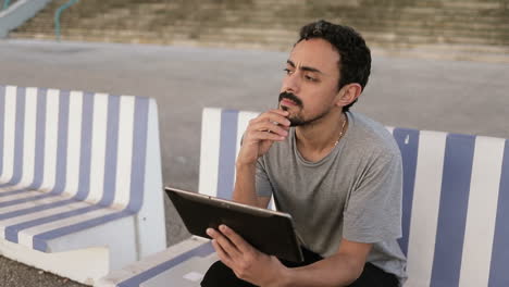Young-Arabic-man-with-dark-curly-hair-and-beard-in-grey-T-shirt-sitting-on-quay-on-striped-bench